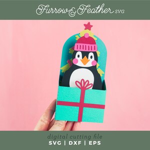 May include: A teal blue gift box with a red ribbon and a penguin wearing a pink hat peeking out of the top. The penguin is surrounded by a green Christmas tree.
