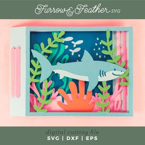 Puede incluir: Una pieza de arte en papel en capas que presenta un tibur&oacute;n azul claro nadando en una escena oce&aacute;nica azul con coral y algas marinas. La obra de arte est&aacute; enmarcada en una caja azul claro. El texto "Furrow & Feather SVG" y "digital cutting file" son visibles.