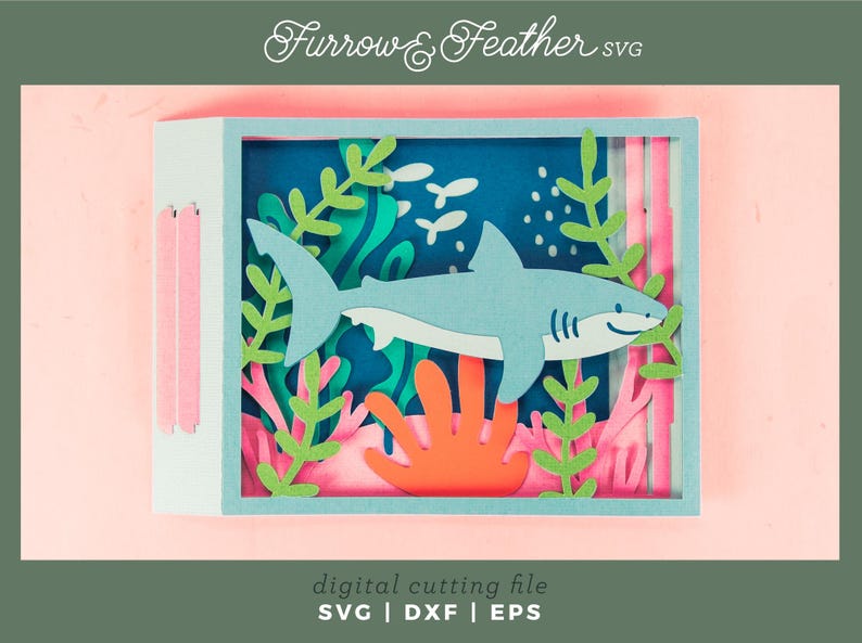 Puede incluir: Una pieza de arte en papel en capas que presenta un tibur&oacute;n azul claro nadando en una escena oce&aacute;nica azul con coral y algas marinas. La obra de arte est&aacute; enmarcada en una caja azul claro. El texto "Furrow & Feather SVG" y "digital cutting file" son visibles.