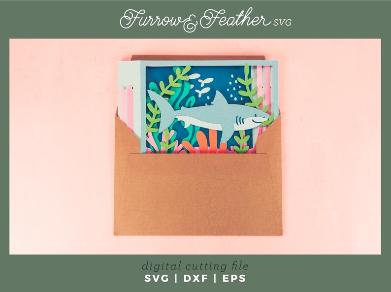Puede incluir: Una escena de tibur&oacute;n de papel en un sobre marr&oacute;n. La escena presenta un tibur&oacute;n azul claro nadando entre coloridos corales y algas marinas. El fondo es azul oscuro. El texto "Furrow & Feather SVG" y "digital cutting file SVG | DXF | EPS" son visibles.