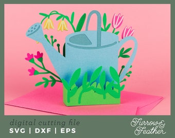 Puede incluir: Una regadera de papel azul claro con hierba verde y flores coloridas. La regadera está rodeada de flores rosas y amarillas y hojas verdes. La imagen incluye el texto "digital cutting file" y "SVG | DXF | EPS".