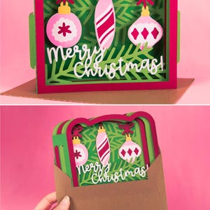 May include: A layered paper Christmas card with a green and red frame, featuring three hanging ornaments and the words "Merry Christmas." The ornaments are pink and white, with various designs. The card is displayed on a pink background.