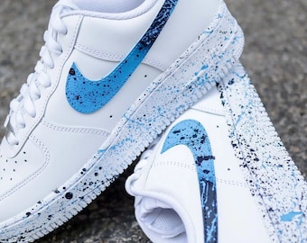 May include: White leather sneakers with a blue and black swoosh logo. The sneakers have a white sole with blue and black paint splatters.