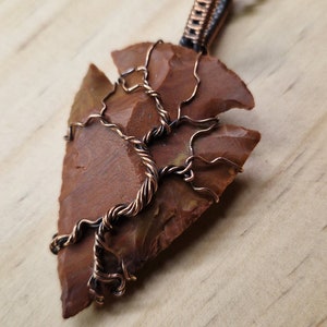 May include: A handcrafted pendant necklace featuring a reddish-brown arrowhead-shaped stone. The stone is intricately wrapped with copper wire, forming a tree-like design. The necklace has a black beaded chain.