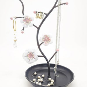 May include: A black metal jewellery stand shaped like a tree with pink and white blossoms. The stand has a round base and branches that curve upwards. There are several necklaces and earrings hanging from the branches.