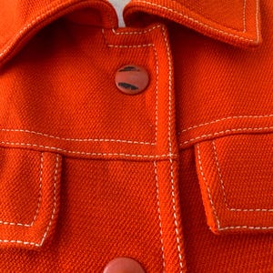 May include: Close-up of a bright orange jacket with white stitching and two round buttons. The jacket has a collar and two front pockets. The stitching is visible along the seams and around the pockets.