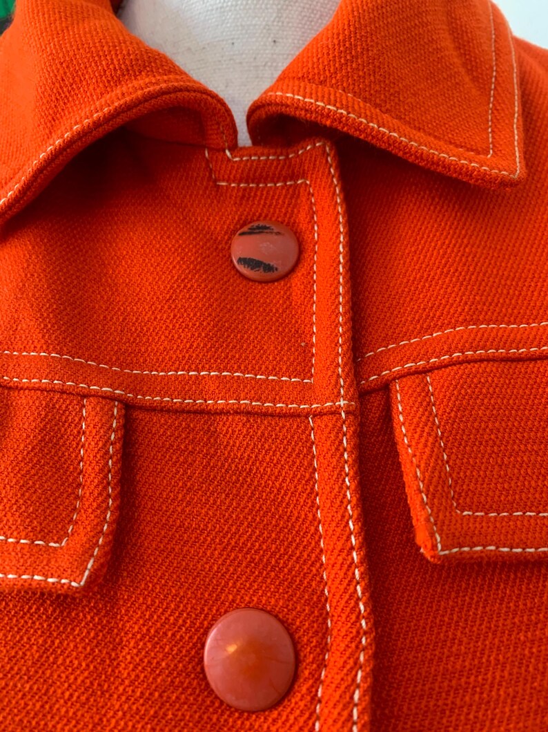 May include: Close-up of a bright orange jacket with white stitching and two round buttons. The jacket has a collar and two front pockets. The stitching is visible along the seams and around the pockets.