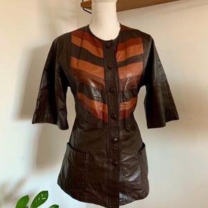 Op de afbeelding: Een vintage bruine leren jas met een uniek ontwerp. De jas heeft horizontale strepen in bruin- en oranjetinten over de borst. Het heeft korte mouwen, een knoopsluiting en twee voorzakken. De jas wordt op een mannequin gepresenteerd.