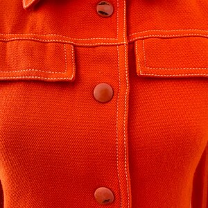 May include: Close-up of a bright orange jacket with a collar, two front pockets, and three matching buttons. The jacket features white stitching along the seams and around the pockets, creating a contrasting detail. The fabric has a textured appearance.