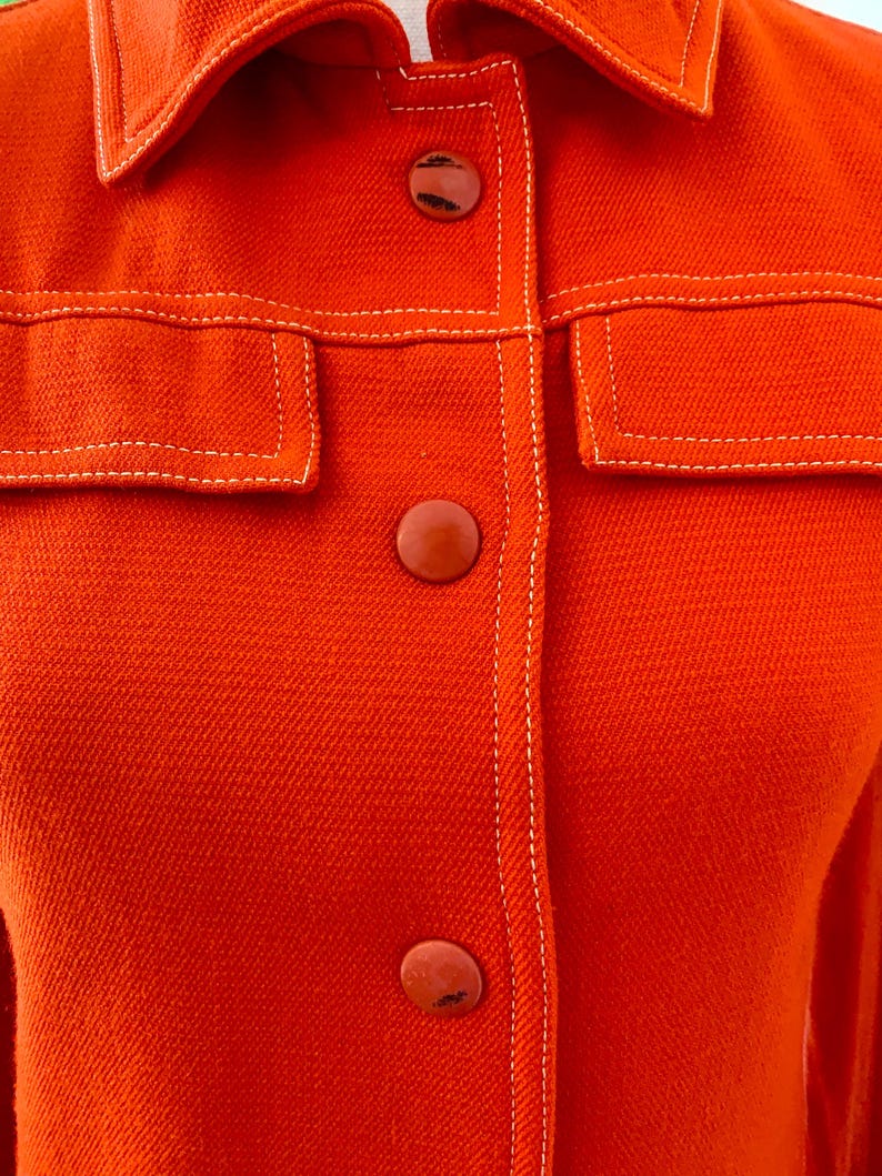 May include: Close-up of a bright orange jacket with a collar, two front pockets, and three matching buttons. The jacket features white stitching along the seams and around the pockets, creating a contrasting detail. The fabric has a textured appearance.