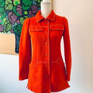 May include: A bright orange jacket with long sleeves, a collar, and button closures. The jacket features two chest pockets and two lower pockets, all outlined with white stitching. The jacket is displayed on a mannequin.