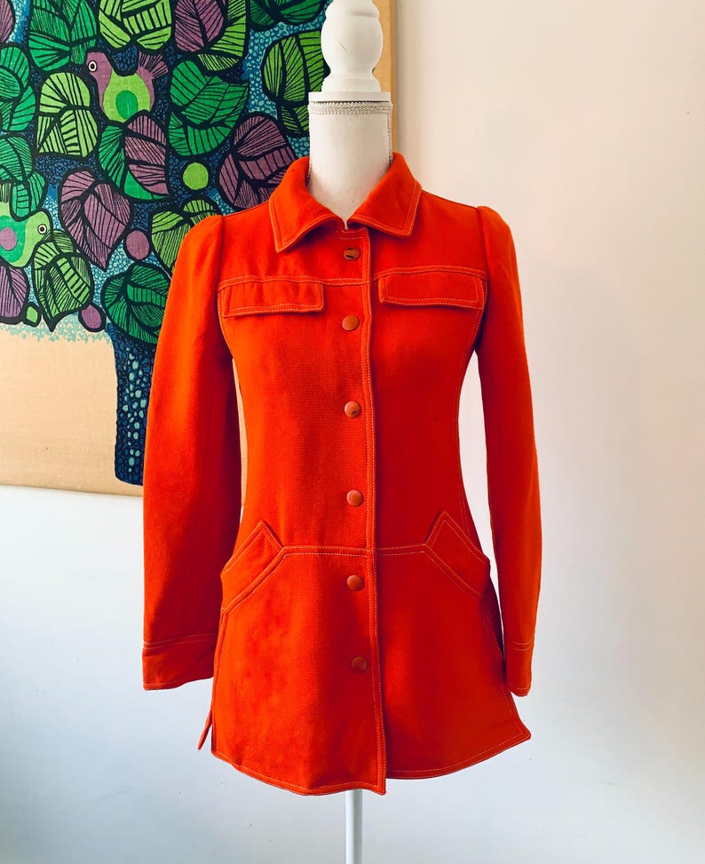 May include: A bright orange jacket with long sleeves, a collar, and button closures. The jacket features two chest pockets and two lower pockets, all outlined with white stitching. The jacket is displayed on a mannequin.