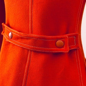 May include: Close-up of a vibrant orange jacket with a tailored fit. The jacket features a belt with two brown buttons and white stitching details. The fabric has a textured appearance, and the sleeves are visible on either side.