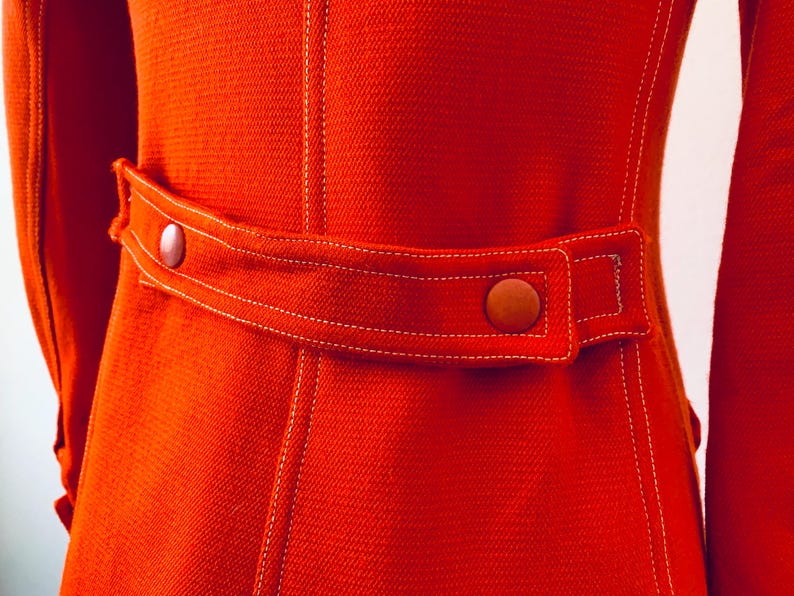 May include: Close-up of a vibrant orange jacket with a tailored fit. The jacket features a belt with two brown buttons and white stitching details. The fabric has a textured appearance, and the sleeves are visible on either side.