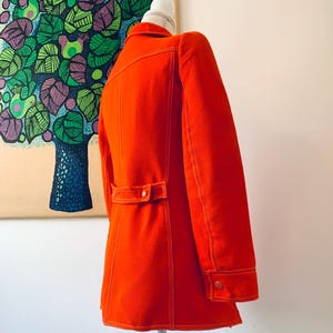 May include: Bright orange vintage jacket with white stitching details. The jacket features a button closure, a notched collar, and a tailored fit. The sleeves have buttoned cuffs. The jacket is displayed on a mannequin.