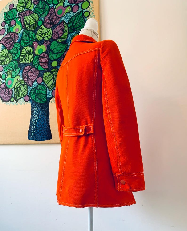 May include: Bright orange vintage jacket with white stitching details. The jacket features a button closure, a notched collar, and a tailored fit. The sleeves have buttoned cuffs. The jacket is displayed on a mannequin.