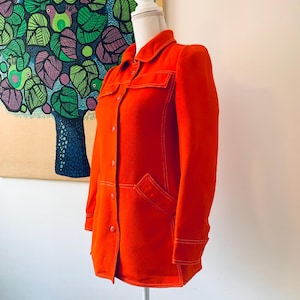 May include: Bright orange jacket with a collared neckline, button-down front, and two chest pockets. The jacket features white stitching details and is displayed on a mannequin. The jacket is a vibrant, solid color.