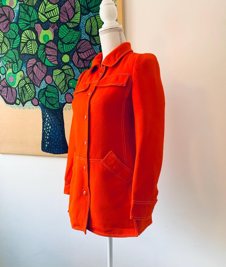 May include: Bright orange jacket with a collared neckline, button-down front, and two chest pockets. The jacket features white stitching details and is displayed on a mannequin. The jacket is a vibrant, solid color.