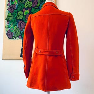 May include: A bright orange vintage coat with white stitching details. The coat features a collar, long sleeves, and a belt detail at the waist. The coat is displayed on a mannequin.