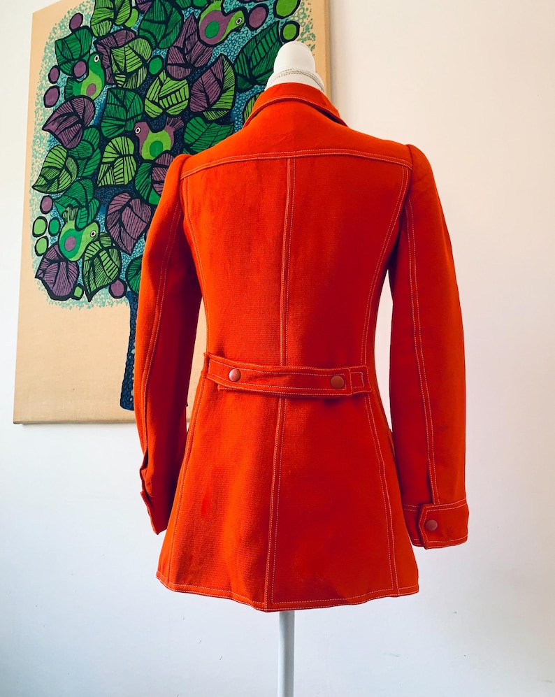 May include: A bright orange vintage coat with white stitching details. The coat features a collar, long sleeves, and a belt detail at the waist. The coat is displayed on a mannequin.