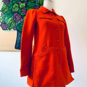 May include: Bright orange vintage coat with a collared neckline, button closures, and large pockets. The coat features white stitching details and is displayed on a mannequin. The coat's design is reminiscent of the 1970s era.