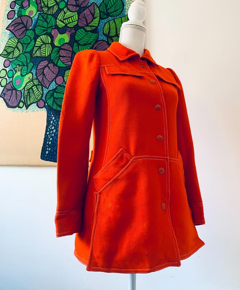 May include: Bright orange vintage coat with a collared neckline, button closures, and large pockets. The coat features white stitching details and is displayed on a mannequin. The coat's design is reminiscent of the 1970s era.