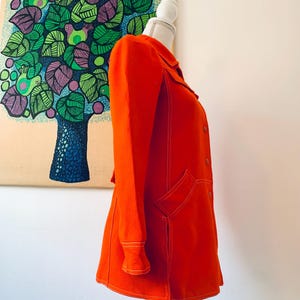 May include: Bright orange coat with white stitching details, displayed on a mannequin. The coat features a collar, long sleeves, and two front pockets. The coat is a vibrant color and has a retro style.