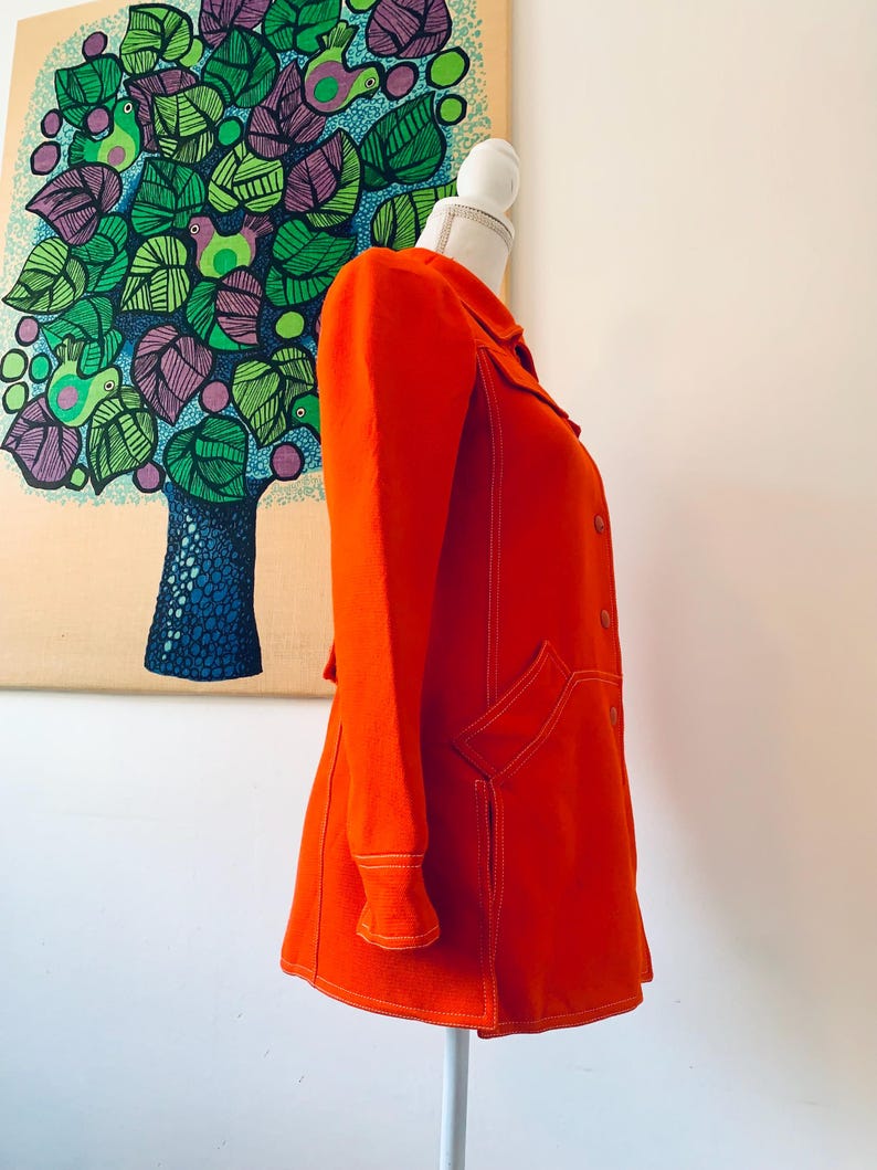 May include: Bright orange coat with white stitching details, displayed on a mannequin. The coat features a collar, long sleeves, and two front pockets. The coat is a vibrant color and has a retro style.