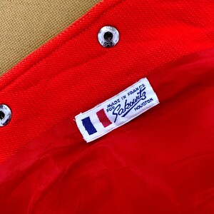 May include: Close-up of a red fabric with a label that reads "MADE IN FRANCE FOR Sakowitz HOUSTON". The label has a small French flag design. The fabric is secured with silver metal snaps.