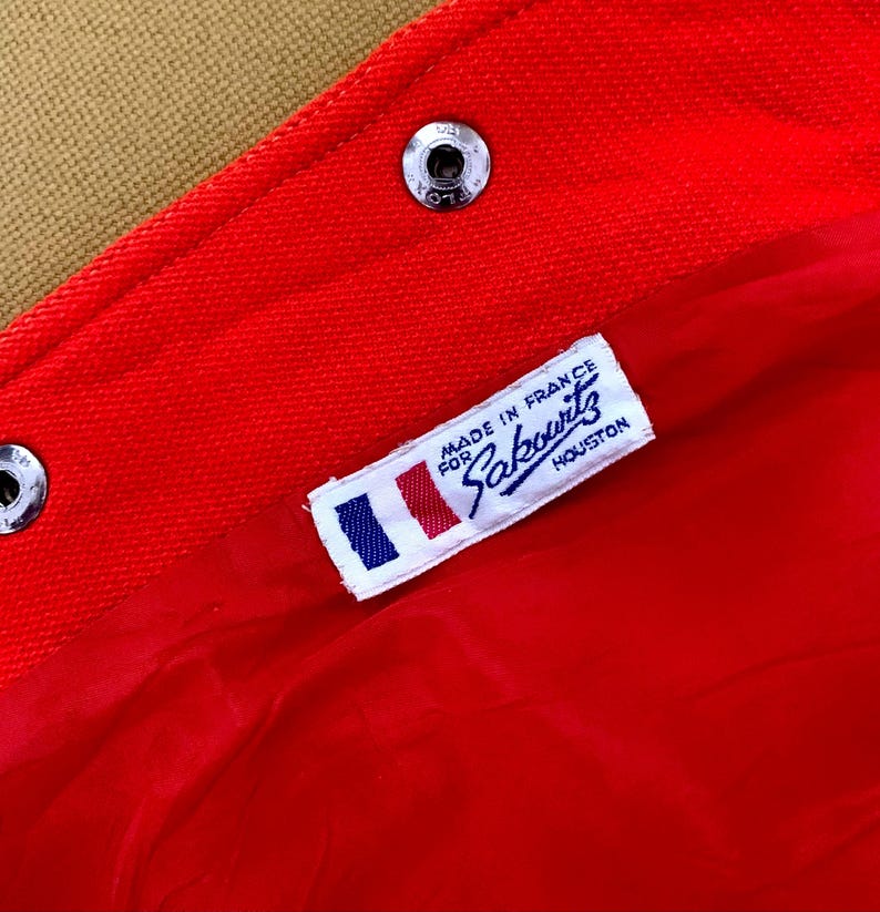 May include: Close-up of a red fabric with a label that reads "MADE IN FRANCE FOR Sakowitz HOUSTON". The label has a small French flag design. The fabric is secured with silver metal snaps.
