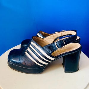 May include: A pair of vintage platform clogs. The shoes are black with white stripes across the top and a buckle strap. The heels are thick and black, and the soles are a dark color.