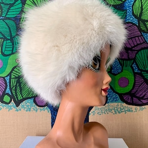 May include: A vintage white fur hat, likely from the mid-20th century, is displayed on a mannequin head. The hat has a rounded shape and a fluffy texture. The mannequin is set against a colourful, patterned backdrop.