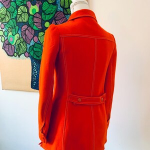 May include: A bright orange jacket with white stitching details, displayed on a mannequin. The jacket features a collar, long sleeves, and a belt detail at the waist. The back of the jacket is visible, showcasing the stitching and design.