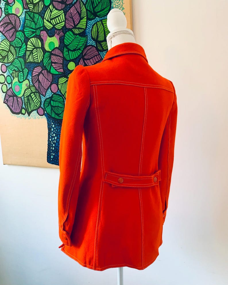 May include: A bright orange jacket with white stitching details, displayed on a mannequin. The jacket features a collar, long sleeves, and a belt detail at the waist. The back of the jacket is visible, showcasing the stitching and design.