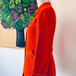 May include: Bright orange jacket with white stitching details, displayed on a mannequin. The jacket features a collar, pockets, and buttoned cuffs. The background includes a colorful tree painting.