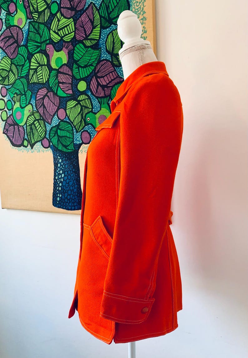 May include: Bright orange jacket with white stitching details, displayed on a mannequin. The jacket features a collar, pockets, and buttoned cuffs. The background includes a colorful tree painting.