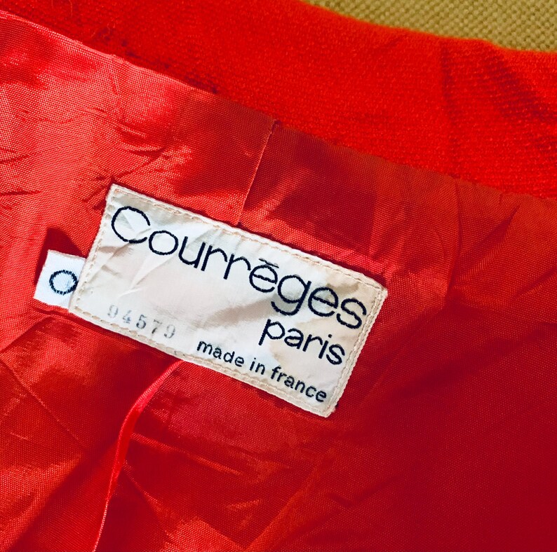 May include: Close-up of a bright red garment interior with a satin lining. A white label reads "Courr&egrave;ges paris made in france" with a small circular tag. The garment's exterior is a textured, solid red.