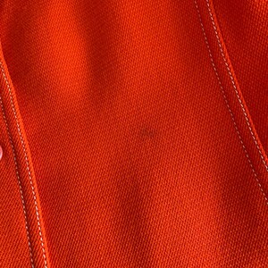 May include: Close-up of a vibrant orange fabric with white stitching. The stitching outlines the seams of a garment, possibly a jacket or coat. The texture of the fabric is visible, suggesting a woven material.