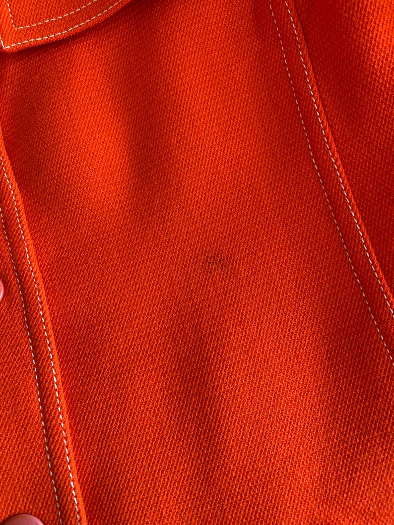 May include: Close-up of a vibrant orange fabric with white stitching. The stitching outlines the seams of a garment, possibly a jacket or coat. The texture of the fabric is visible, suggesting a woven material.
