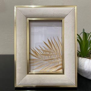 May include: A rectangular photo frame with a gold trim and a cream velvet border. The frame displays a picture of a golden palm leaf. The frame has the text "Neutral Gold Edged Velvet Frame".