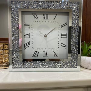 May include: Square mirrored clock with a silver crystal-studded frame. The clock face is white with black Roman numerals and hands. A decorative accent for a living room or bedroom.