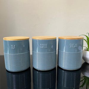 May include: Three matching kitchen storage canisters in a muted blue-grey. Each has a ribbed lower section and a wooden lid. The canisters are labelled "TEA", "COFFEE", and "SUGAR" in embossed white lettering. Ideal for tea, coffee, and sugar.