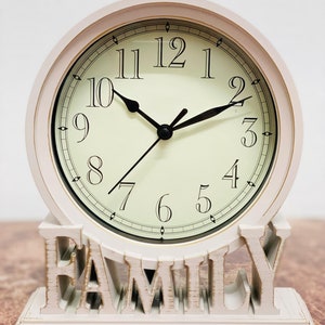 May include: White tabletop clock with a round face and black hands. The clock face has black Roman numeral markings and a gold trim. The clock is sitting on a white base with the word "FAMILY" spelled out in large letters.