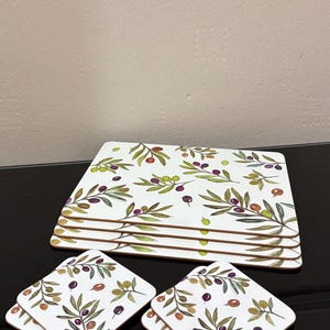 May include: A collection of white ceramic plates and coasters with an olive branch design. The plates are rectangular, and the coasters are square. The design features green leaves, purple olives, and brown stems. Ideal for serving food and drinks at home.