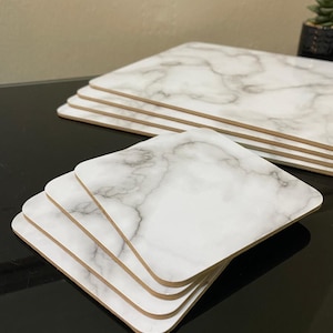 May include: A collection of marble-effect coasters and placemats. The coasters are square with rounded edges, and the placemats are rectangular. The white and grey marble pattern contrasts with the wooden edges. The items are stacked on a black surface.