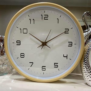 May include: A round, cream-coloured wall clock with black numbers and hands. The clock face is white and has a light blue outer ring.