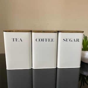 May include: Three rectangular kitchen storage canisters. The top half of each is white, with the words "TEA", "COFFEE", and "SUGAR" printed in grey. The bottom half is grey. Each canister has a wooden lid.