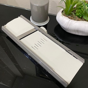 May include: A white and grey mandoline slicer with a stainless steel blade, next to a grey container and a small potted plant. The slicer has a ribbed surface and adjustable settings for various cuts. Ideal for slicing vegetables and fruits.