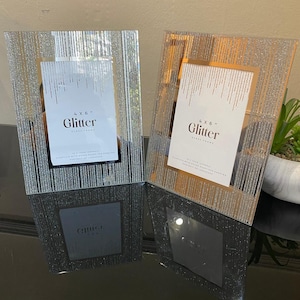 May include: Two glass photo frames with glitter detailing. Each frame holds a 10x15 cm photo and features the word "Glitter" on the insert. The frames have a vertical striped design and are displayed on a reflective surface.