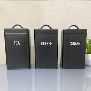 May include: Three black metal canisters with white lettering. The canisters are labelled "TEA", "COFFEE", and "SUGAR".
